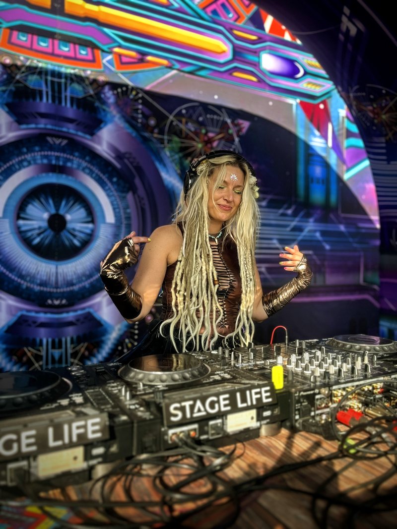 Djane Anastazja smiling behind the decks, long blonde dreadlocks, in front of a psychedelic backdrop with a giant eye