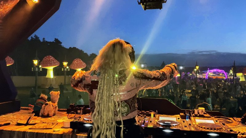 Djane Anastazja seen from behind at the decks, arms out, playing to a festival crowd at dusk