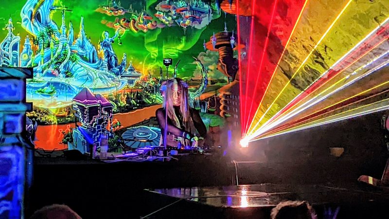 Djane Anastazja playing on a stage with a bright psychedelic backdrop and fans of laser light