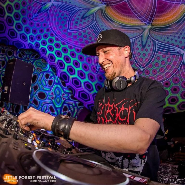 DJ Archive grinning at the decks under a purple and green UV backdrop at an Alien Safari festival, Technics headphones around his neck.
