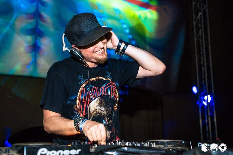 DJ Archive at the decks in a Slayer T-shirt and cap, headphones on, grinning under blue and green stage lighting.