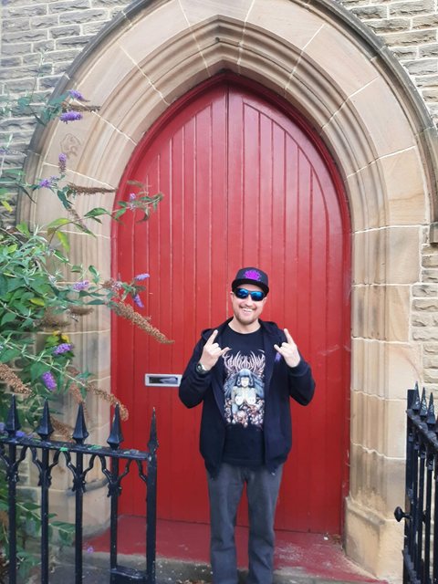 DJ Archive throwing metal horns in sunglasses and a death-metal T-shirt, in front of a red gothic arched door.
