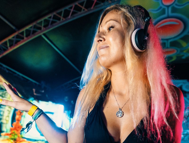 Djane Anastazja at the decks, headphones on, long blonde and pink hair, under a colourful psychedelic backdrop