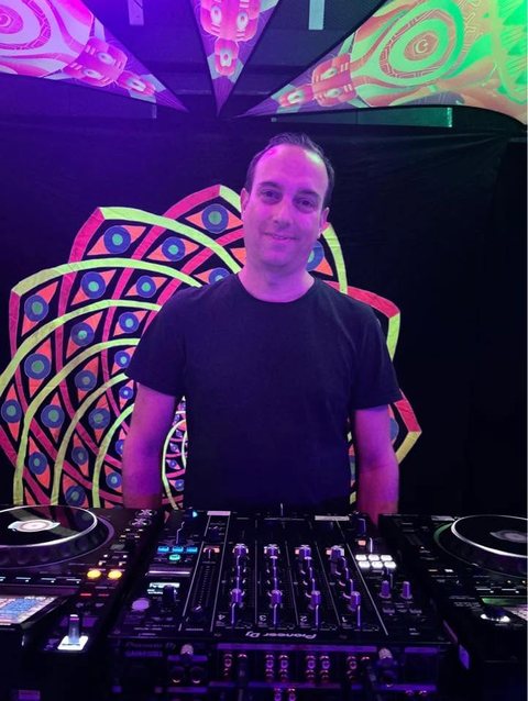 Aurel behind the decks at a psytrance party, smiling, with a UV mandala backdrop and psychedelic banners overhead.