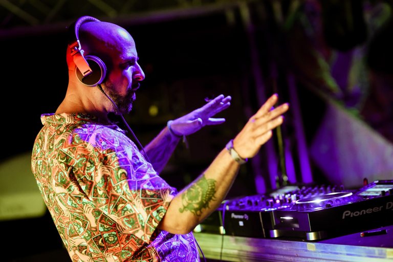 Champa at the decks, headphones on, both hands raised to the crowd under purple light