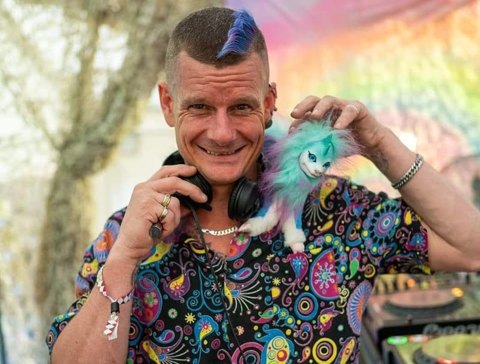 DJ S&M smiling at the decks, headphones on, a blue streak in his hair and a small plush creature on his shoulder