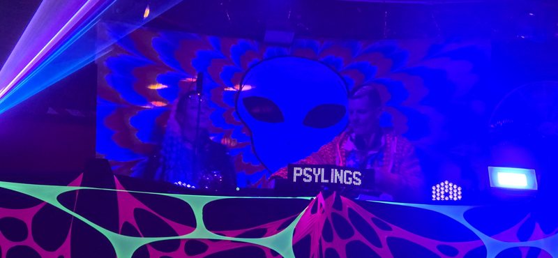 The Psylings in the DJ booth under a giant UV alien backdrop, a PSYLINGS sign on the decks