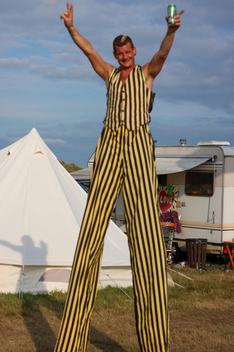 DJ S&M on stilts in yellow-and-black striped trousers and waistcoat, arms raised, at a festival campsite with a bell tent and a caravan behind