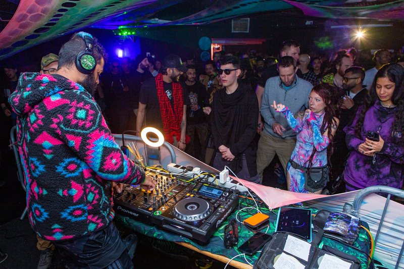 R3jal from behind in a patterned fleece and R3JAL headphones, mixing on Pioneer decks, a club crowd close in front of the booth.