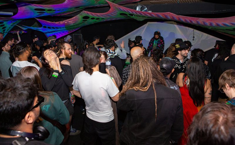 A packed club floor under UV canopy drapes, people dancing, R3jal at the decks at the back of the room.