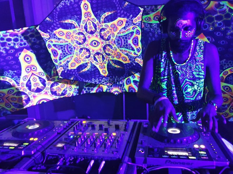 R3jal at silver Pioneer decks under UV light, face paint glowing, fluorescent psychedelic backdrops behind him.