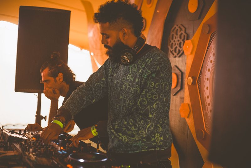 R3jal mixing in warm tent light, eyes down, R3JAL headphones round his neck, another DJ leaning on the booth beside him.