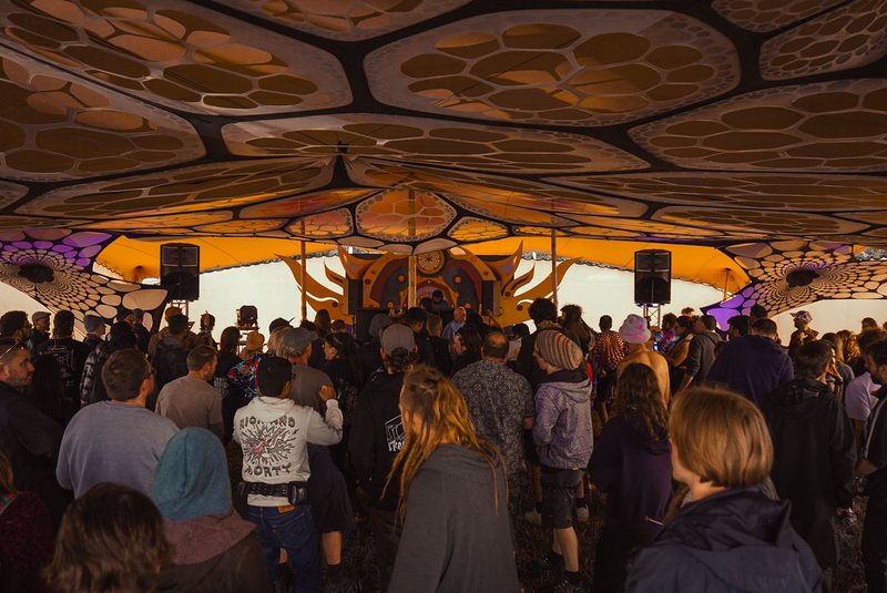 A crowd under a patterned festival canopy facing a decorated stage, R3jal small at the decks in the distance.