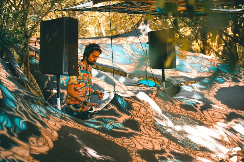 R3jal at the decks, headphones on, on an outdoor forest stage draped in psychedelic fabric between two speakers.