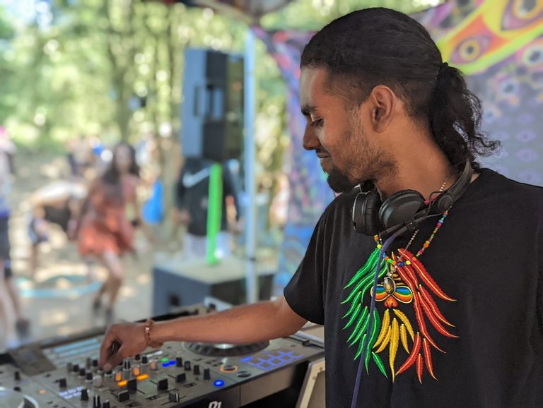 R3jal smiling at the mixer on a daytime outdoor stage, headphones around his neck, dancers behind him among the trees.