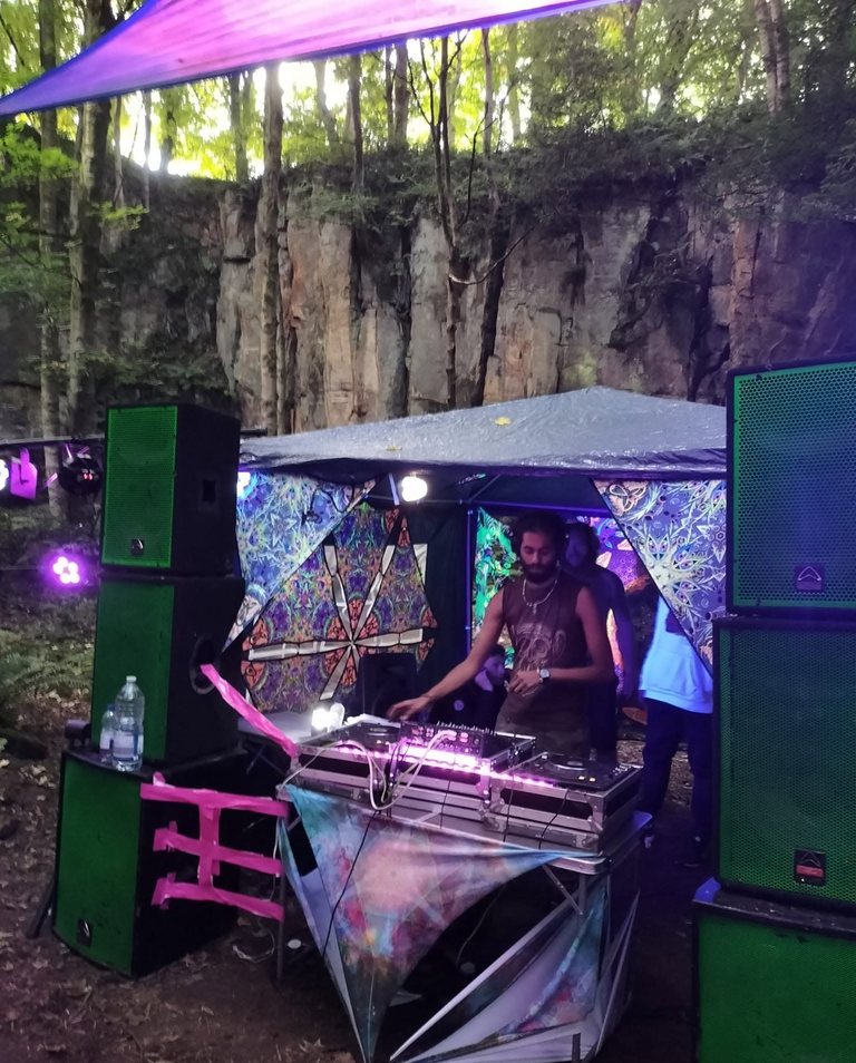 R3jal at the decks under a gazebo at an outdoor party in a woodland quarry, stacked green speakers either side and a rock face behind.