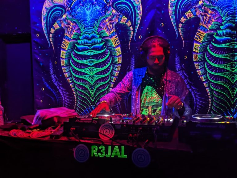 R3jal at the decks under UV light, headphones on, in front of glowing psychedelic backdrops, his name lit up on the front of the booth.