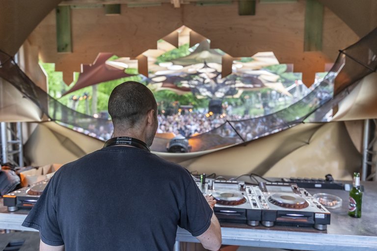 Sabretooth seen from behind the decks, looking out through a geometric stage canopy at a packed daytime festival crowd.