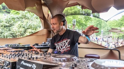 Sabretooth (Ben Fraser) at the decks in a daytime festival booth, headphones on, one hand raised, green foliage behind him.