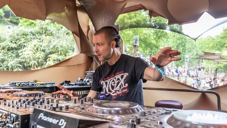 Sabretooth (Ben Fraser) at the decks in a daytime festival booth, headphones on, one hand raised, green foliage behind him.
