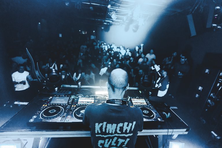 Sabretooth from behind the decks under a spotlight, facing a packed club crowd lit in deep blue.